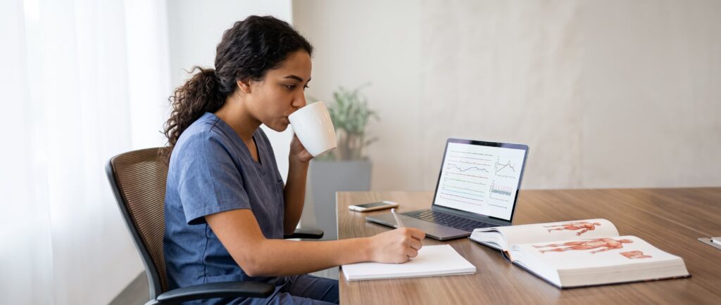 Woman in scrubs drinks from a mug, writes notes at desk with laptop and open book displaying medical diagrams.