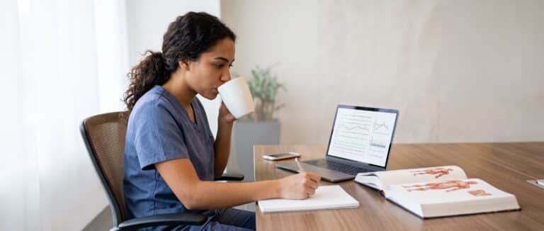 Woman in scrubs drinks from a mug, writes notes at desk with laptop and open book displaying medical diagrams.